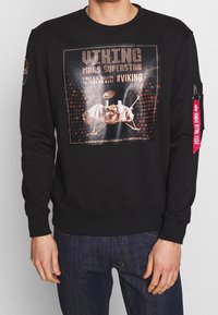 Persona vestida con un sudadera negra que tiene un gráfico del módulo de aterrizaje Viking de Marte y un parche del logo de la NASA en la manga, acompañada de unos vaqueros azules.