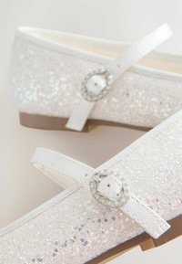 Ballet flats bianche e glitterate con punta curvata, una strap decorativa con una fibbia rotonda e una suola in gomma marrone.