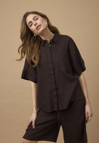Vrouw met lang bruin haar, gekleed in een losse donkerbruine korte mouwen shirt en bijpassende korte broek, poserend tegen een beige achtergrond.
