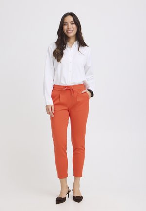 Frau steht, lächelt, trägt ein weißes, langärmeliges Hemd mit Knöpfen, orangefarbene, maßgeschneiderte Hosen mit Kordelzug und schwarze spitze High Heels.