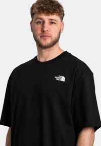 T-shirt in cotone nero con vestibilità rilassata, caratterizzato da un logo bianco sul petto sinistro e maniche corte. Texture liscia, design semplice.