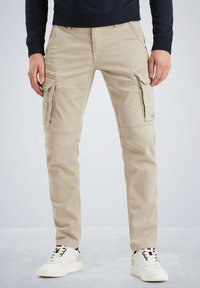 Beige cargo bukser med flere sidelommer, slim fit og glat bomuldsstof. Båret med hvide sneakers, der har sorte snørebånd.