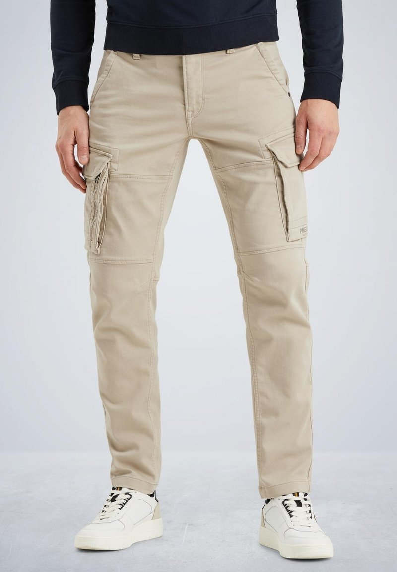 Beige cargo bukser med flere sidelommer, slim fit og glat bomuldsstof. Båret med hvide sneakers, der har sorte snørebånd.