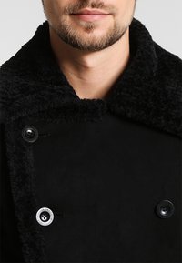 Manteau noir avec col en peau lainée, doté de deux boutons ronds et d'une texture lisse. Vue rapprochée mettant en valeur les détails du design.