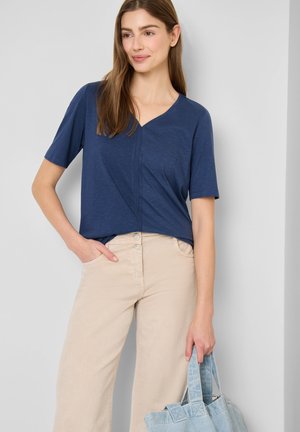 Kvinde iført en mørkeblå V-neck bluse og beige bukser, der holder en lyseblå stoftaske med højre hånd.