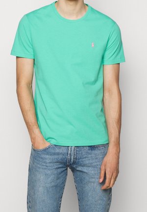 Mężczyzna noszący miętowo-zieloną koszulkę z krótkim rękawem i małym różowym logo na piersi, w połączeniu z jasnoniebieskimi dżinsami, jedną ręką w kieszeni.