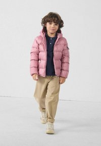 Chaqueta acolchada rosa claro con capucha, bolsillos laterales y costuras visibles, combinada con una camiseta oscura y pantalones beige, en un niño.
