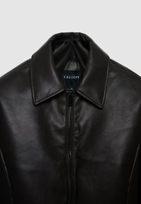 Veste en cuir noir avec un col, dotée d'une fermeture éclair centrale. Le matériau a une texture lisse et peu de détails. Étiquette de marque visible.