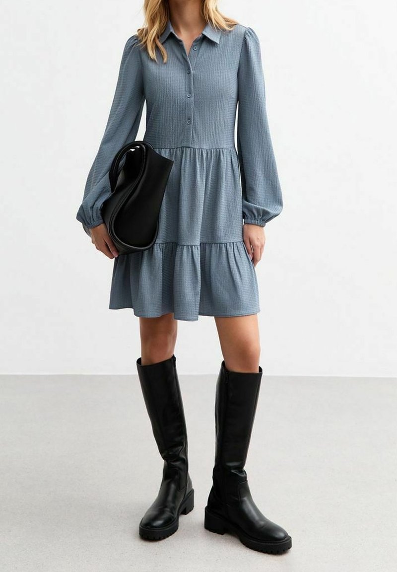 Femme portant une robe bleue à manches longues boutonnée, tenant un sac à main noir et portant des bottes noires montantes, sur un fond uni.