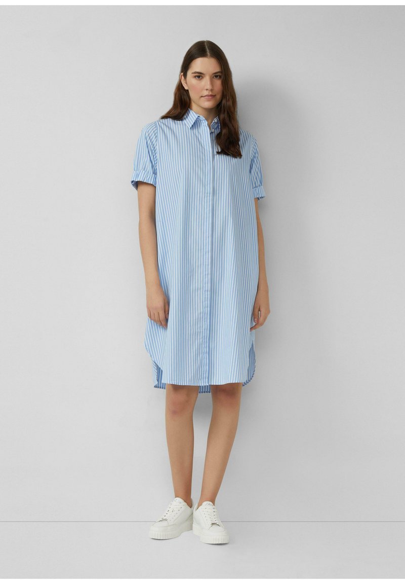 Donna che indossa un abito camicia blu e bianco a righe verticali, lungo fino al ginocchio, con maniche corte arrotolate e sneakers bianche.
