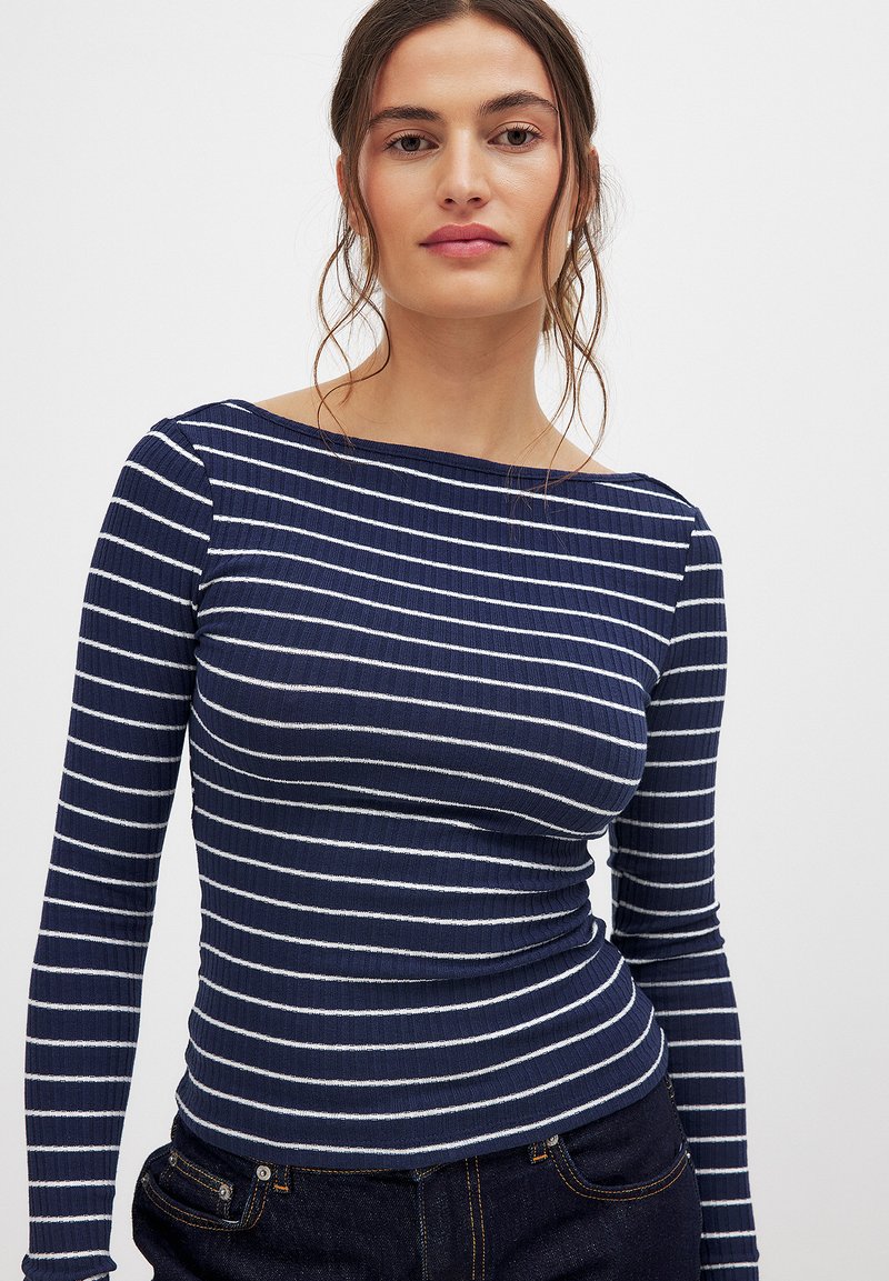 Marineblaues Langarmoberteil mit weißen horizontalen Streifen, ausgestattet mit einem Bootsausschnitt und gerippter Textur. Kombiniert mit dunklen Jeans.