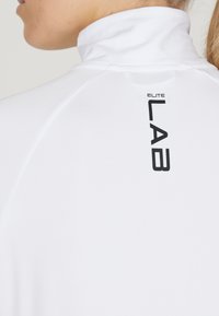 Weißes Sportoberteil mit hohem Kragen. Es hat einen strukturierten Stoff und ein schwarzes Logo "ELITE LAB", das auf dem Rücken aufgedruckt ist.