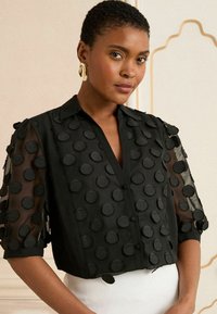 Čierna blúzka s priesvitnými rukávmi, so veľkými textúrovanými bodkami. Okrúhly golier a zapínanie na gombíky s oblým spodným lemom.