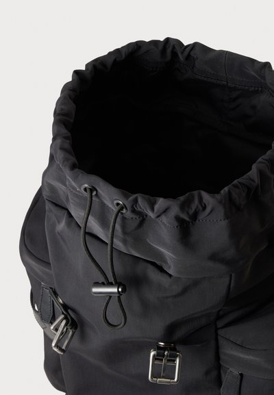 Sac à dos noir avec ouverture à cordon en haut, deux poches latérales et des fermetures à boucle en métal, vu de dessus sur un fond clair.
