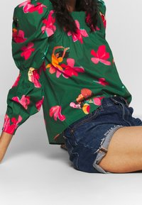 Blouse verte à motif floral rose et jaune, manches bouffantes et encolure smockée. Associée à un short en denim aux bords effilochés.