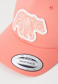 Koralā krāsā snapback cepure ar baltu lāča uzšuvi, kas izceļas ar sarežģītu līniju rakstu. Iekļauts sudraba apļa uzlīme ar zīmola detaļām.