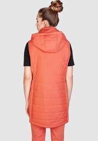 Oranger ärmelloser Steppweste mit Kapuze, mit einer gesteppten Textur und einem längeren Schnitt, getragen über einem schwarzen Shirt.