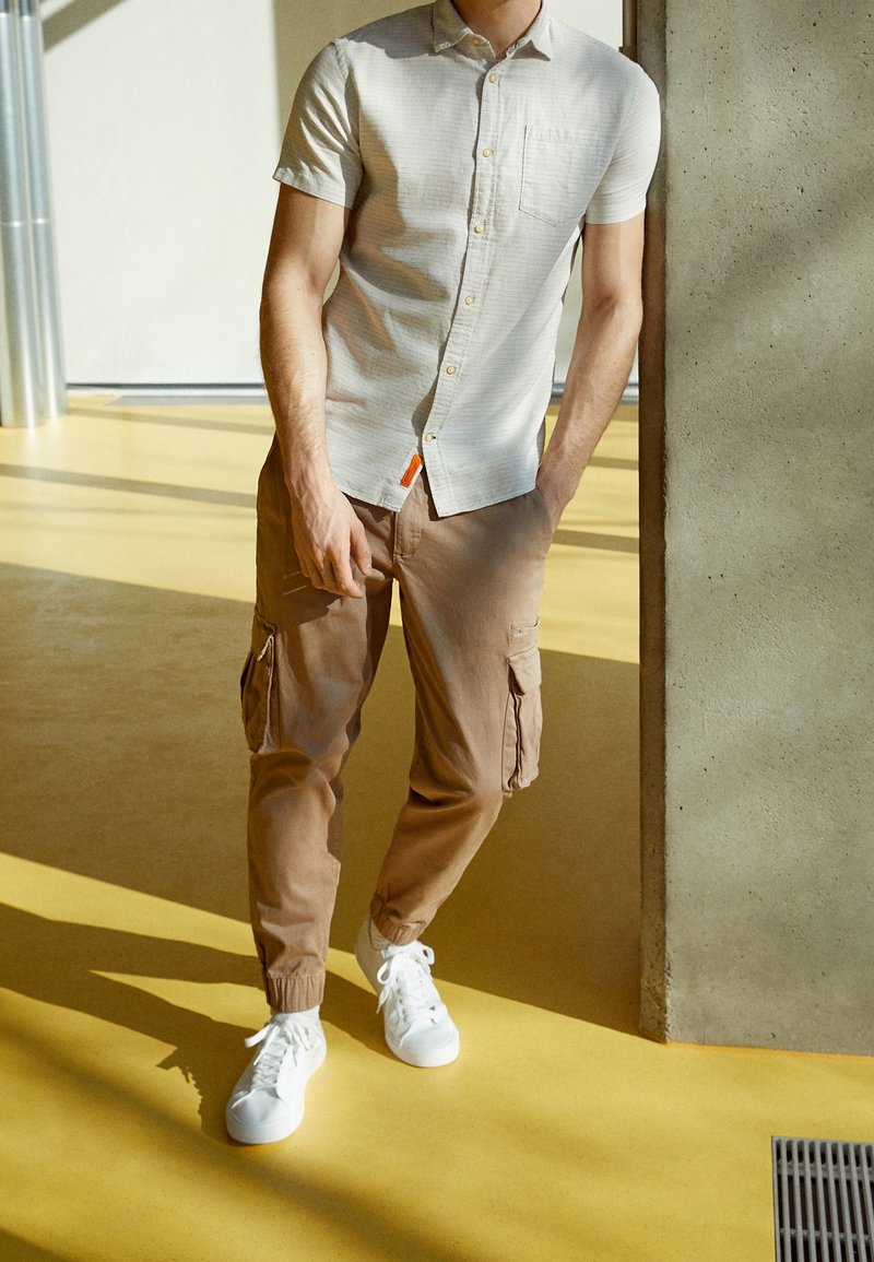 Homme portant une chemise à manches courtes claire à boutons, un pantalon cargo marron et des baskets blanches, debout à l'intérieur près d'un pilier en béton.