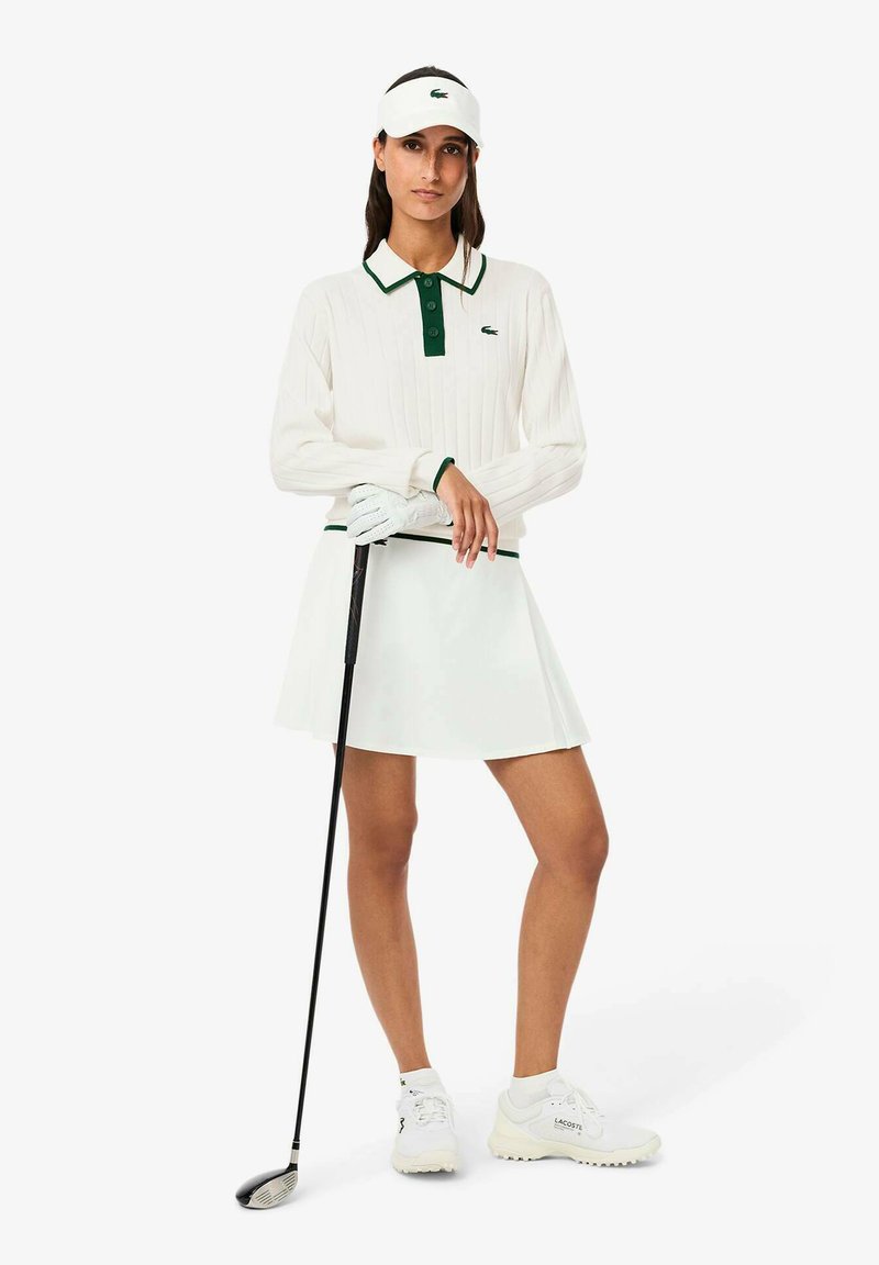 Witte geribbelde polo met groene kraag en knopen, gecombineerd met een witte rok. Accessoires omvatten een wit vizier en een golfclub, met witte sneakers.