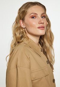 Beige Knopfjacke mit Kragen, ausgestattet mit einer Brieftasche auf der Vorderseite und einer glatten Textur. Die Haare sind in sanften Wellen gestylt.
