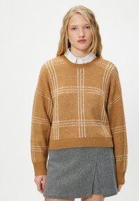 Senfgelbes gestricktes Sweater mit weißem Karomuster, rundem Halsausschnitt und gerippten Bündchen. Kombiniert mit einem grauen plissierten Minirock.