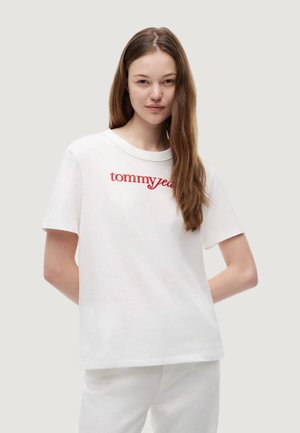 Jeune femme aux longs cheveux bruns portant un t-shirt blanc avec un texte rouge "tommyjeans" et un pantalon blanc sur un fond uni.