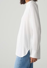 Blouse blanche à manches longues avec un col montant, des fentes latérales et des manches à revers. Fabriquée en tissu léger avec une texture lisse.