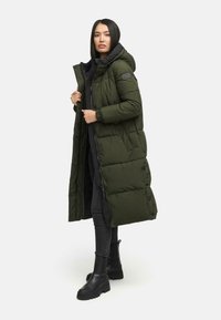 Lang olivengrøn pufferjakke med hætte, der har lynlåslukning, trykknapper og sidelommer, båret over sorte klæder og støvler.