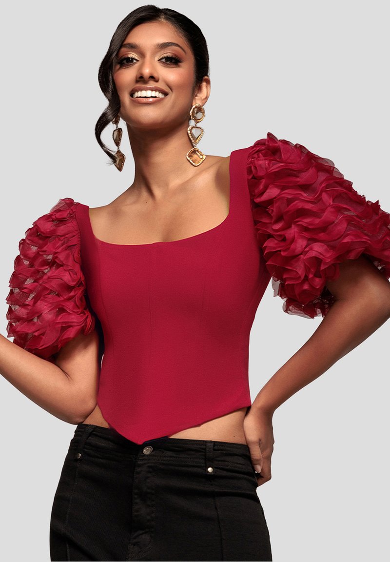 Rød tætsiddende top med firkantet halsudskæring og korte, flæseformede gennemsigtige ærmer, kombineret med sort højtaljede bukser. Glat tekstur.