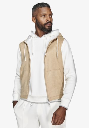 Uomo con barba che indossa felpa con cappuccio bianca e pantaloni con gilet senza maniche beige con zip, che guarda verso sinistra, mani rilassate.