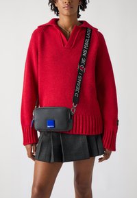 Red knit sweater with a collar and ribbed hem, paired with a black rectangular crossbody bag featuring a blue accent and adjustable strap.