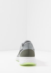 Baskets de sport grises avec détail vert au talon, semelle blanche texturée et semelle extérieure vert clair, présentées de dos sur une surface blanche.