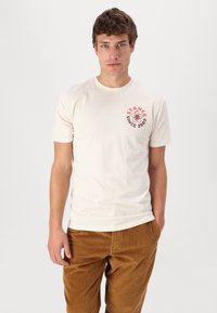 Beige bomulds t-shirt med rund hals og korte ærmer. Grafikken på forsiden viser "STANCE" i rød og et blomsterdesign. Bæres med brune kordfløjl bukser.