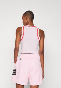 Witte tanktop met rode bies, gecombineerd met roze shorts met zwart drie-strepen logo op de zijkant. De outfit is sportief en strak fitting.