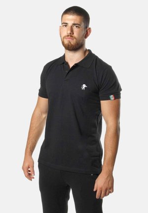 Man met baard die een zwart poloshirt draagt met een wit leeuwenlogo op de borst en een Italiaans vlagpatch op de mouw, staand tegen een witte achtergrond.