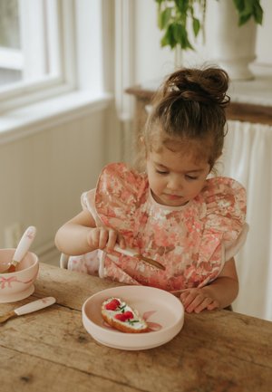 Young girl with hair in a bun spreads topping on bread with a knife at a wooden table, wearing a floral bib and light pink dress.