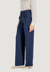 Pantalons navy à jambes larges en tissu lisse, avec des plis, une taille haute et une coupe ajustée. Associés à une chemise légère à manches longues texturée.