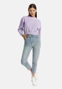 Lila cropped pulóver fehér felirattal, világoskék, magas derekú farmernadrággal és rózsaszín részletekkel ellátott fehér sportcipővel párosítva.