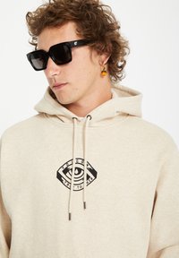 Beige hoodie met zwart grafisch ooglogo, trekkoorden met metalen punten. Model draagt zwarte vierkante zonnebril en een gouden ringoorbel met een oranje bedel.