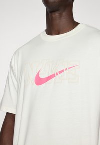 Bílá bavlněná tričko s výrazným růžovým logem Nike a zašlým béžovým písmem. Kulatý výstřih, krátké rukávy, volný střih.