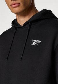 Čierna mikina s kapucňou vyrobená z bavlnenej zmesi, s bielym logom Reebok na hrudi, so sťahovateľnou kapucňou a rebrovanými manžetami.