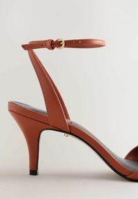 Sandale à talon haut en cuir marron avec bride à la cheville et boucle dorée, semelle noire et talon moyen effilé sur fond neutre.