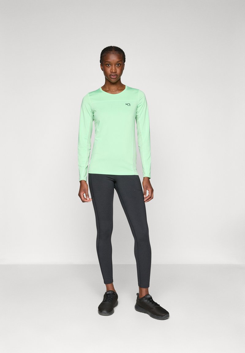 Kobieta stojąca w miętowo-zielonym długim topie, ciemnoszarych legginsach i czarnych butach sportowych na jednolitym jasnym tle.