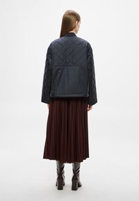 Tengerészkék steppelt bomberdzseki álló gallérral és deréknál vízszintes varrással, bordó pliszírozott szoknyával és térdig érő barna csizmával párosítva.