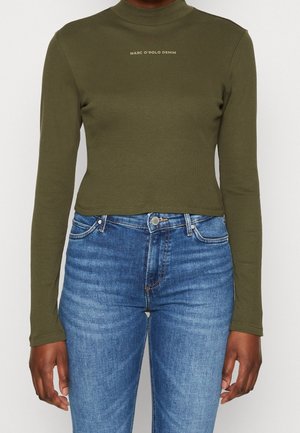 Vrouw draagt een olijfgroen langarmshirt met de tekst "MARC O'POLO DENIM" en een hooggetailleerde blauwe spijkerbroek, staand tegen een effen achtergrond.