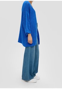 Modrý oversized pletený svetr s výstřihem do V a širokými rukávy, spárovaný s světle modrými širokými džínami a bílými teniskami.