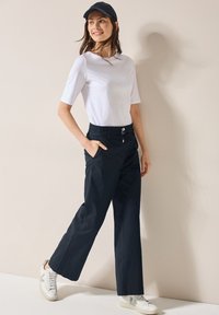 Aansluitend wit katoenen t-shirt, marineblauwe wijde broek met een trekkoord en knoopsluiting, gecombineerd met witte sneakers en een marineblauwe pet.