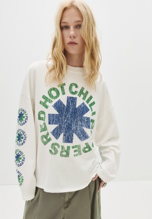 Jongere met lang blond haar, gekleed in een witte lange mouwen shirt met een groen en blauw Red Hot Chili Peppers-logo en olijfgroene broek.