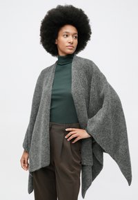 Grå ullwrap med brede ermer, åpen front og myk tekstur. Modellen har på seg en mørkegrønn strikket genser og brune skreddersydde bukser.