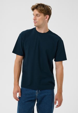Joven con cabello castaño claro que lleva una camiseta de manga corta azul oscuro y vaqueros azules, mirando hacia su izquierda con un fondo liso.